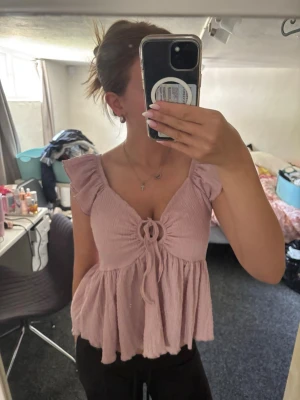 Ljusrosa blus med volang och knyt - Supersöt ljusrosa blus med volangärmar och knyt framtill. Blusen har en v-ringad design och är tillverkad i ett lätt, skrynkligt material som ger en avslappnad känsla. Perfekt att styla med jeans eller kjol för en trendig look.