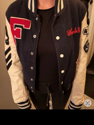 Mörkblå och vit bomberjacka Franklin - Snygg collegeinspirerad bomberjacka i mörkblått med vita ärmar och detaljer. Stora röda bokstäver och broderier med texten Franklin och 99 på ryggen. Jackan har ribbade muddar och tryckknappar framtill. Perfekt för en sportig och retro look.