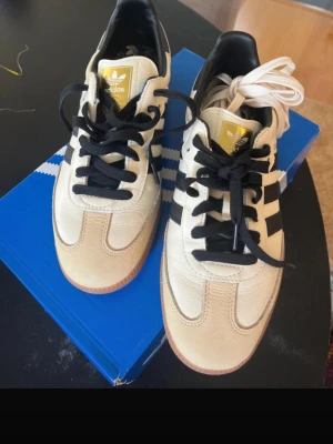 Adidas Samba OG vita och svarta sneakers - Säljer ett par klassiska Adidas Samba OG sneakers i vitt och svart med gummisula. Skorna har svarta ränder på sidorna, svart häl och svart samt vit snörning. Ovandelen är i läder och mocka, med gulddetaljer på plösen. Perfekta för en sportig och stilren look.