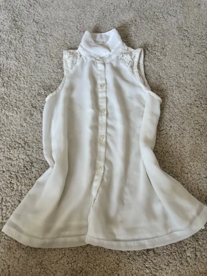 Vit ärmlös blus med spetsdetaljer - Stilren vit blus i ett lätt, flowy material med hög krage och knappar framtill. Ärmlös design och fina spetsdetaljer vid axlarna ger en elegant touch. Perfekt att matcha med jeans eller kjol för en chic look.