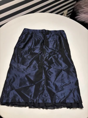 Mörkblå långkjol i satin - Snygg mörkblå långkjol i glansigt satinliknande material med volangkant längst ner. Kjolen har en rak passform och elastisk midja för extra komfort. Perfekt för dig som vill ha en elegant och stilren look.