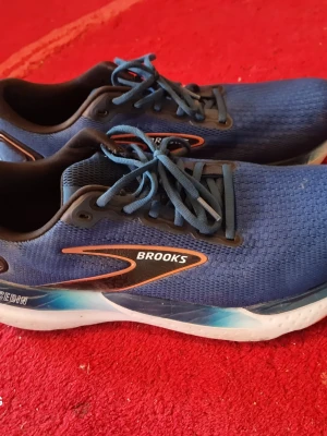 Blå Brooks sneakers med snörning - Snygga blå sneakers från Brooks med svart och orange detalj på sidan. Ovandelen är i meshmaterial för bra ventilation och skorna har klassisk snörning. Sulan är vit med blå inslag och ger en sportig look. Perfekta för dig som gillar stilrena och bekväma sneakers.