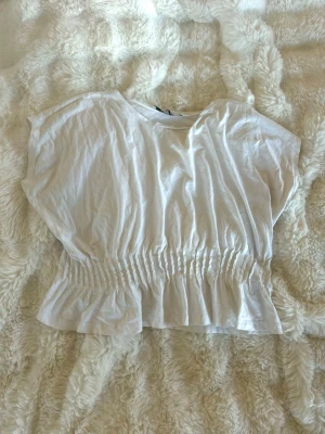 Vit croppad topp med resår i midjan - Säljer en vit croppad topp med korta ärmar och rund hals. Toppen har resår i midjan som ger en snygg rynkad effekt och volang nertill. Perfekt för en chill och trendig look.