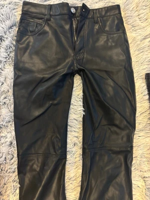 Svarta skinnbyxor från Bershka - Säljer ett par svarta skinnbyxor från Bershka i storlek S. Byxorna har klassisk femficksmodell, dragkedja och knapp framtill samt raka ben. Snyggt glansigt material som ger en cool look till din outfit. sydda för att vara mer lowaist