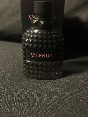 Valentino svart parfymflaska - Snygg parfym från Valentino i en svart, nitar-inspirerad glasflaska med matt finish och rosa text på etiketten. Flaskan har en cool, edgy design som sticker ut och känns lyxig. Perfekt för dig som gillar exklusiva dofter och vill ha en parfym som syns i badrumshyllan.