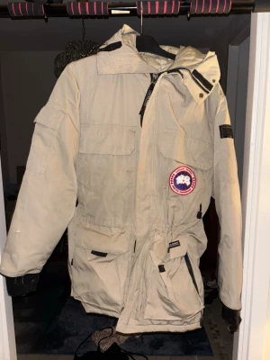Beige canada goose jacka med patch - Snygg beige canada goose jacka med huva och flera praktiska fickor. Jackan har en tydlig patch på bröstet och justerbar midja. Perfekt för kalla dagar med sin långa modell och vindtäta material. Stängs med dragkedja och kardborreband.