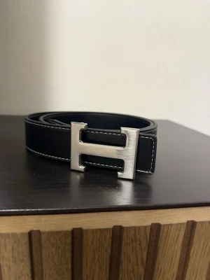 Svart bälte - Snyggt svart bälte. Bältet har vita kontrastsömmar längs kanterna som ger en stilren look. Perfekt accessoar för att lyfta din outfit.