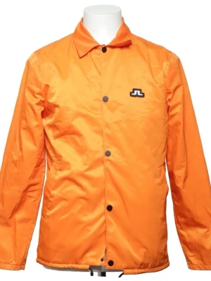 Orange vindjacka med svart logga - Snygg orange vindjacka med svart tryckknappsstängning och stor svart logga på ryggen samt mindre logga på bröstet. Jackan har klassisk krage och dragsko nertill. Perfekt för dig som vill sticka ut med färg och stil.