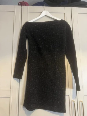 Svart glittrig långärmad klänning - Snygg svart klänning med glittriga detaljer och lång ärm. Modellen är kroppsnära och har en klassisk, rak form som slutar ovanför knäna. Perfekt för dig som vill sticka ut med lite extra glitter. Testad på men aldrig använd