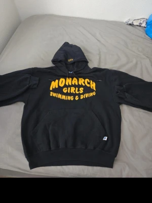 Svart hoodie Monarch Girls - Svart hoodie med gul text 'MONARCH GIRLS SWIMMING & DIVING' på bröstet. Klassisk känguruficka framtill och huva med dragsko. Tillverkad i mjukt material, perfekt för chill dagar eller träning.
