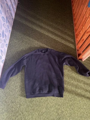 Svart basic sweatshirt - En enkel svart sweatshirt med rund hals och långa ärmar. Tröjan har ribbade muddar vid ärmslut och nederkant. Perfekt för en clean och avslappnad stil. Materialet ser ut att vara mjukt och bekvämt, troligen bomull eller bomullsblandning.