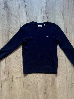 Mörkblå stickad tröja från GANT - Mörkblå ullstickad tröja från GANT med rund halsringning och diskret broderad logga på bröstet. Tröjan har ribbade muddar vid ärmslut och nederkant. Perfekt för en stilren och klassisk look.