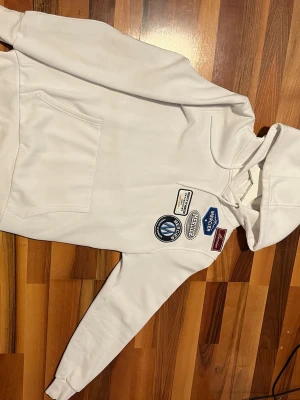 Vit hoodie med patchar och huva - Snygg vit hoodie med flera patchar och tryck på bröstet. Klassisk känguruficka framtill och dragsko i huvan. Tillverkad i mjukt material som känns skönt mot huden. Perfekt för dig som gillar streetwear och vill sticka ut lite extra.