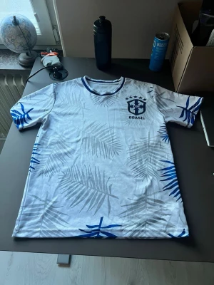 Vit Brasilien fotbollströja med blå detaljer - Säljer en vit fotbollströja med Brasilien-emblem på bröstet. Tröjan har blå detaljer och palmbladsmönster över hela tyget samt blå kant vid halsen. Perfekt för dig som vill sticka ut på planen eller heja på Brasilien.
