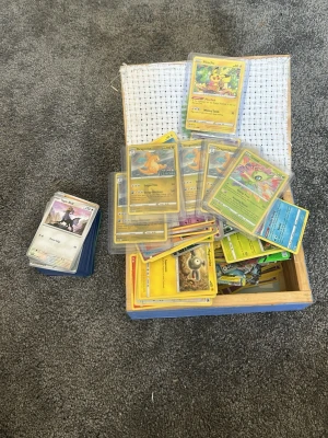 Stor samling Pokémonkort i trälåda - Säljer en stor samling Pokémonkort i olika färger och motiv, förvarade i en blå trälåda. Flera kort är i plastfickor och inkluderar populära karaktärer som Pikachu och Dragonite. Perfekt för samlare eller dig som vill utöka din kortlek.