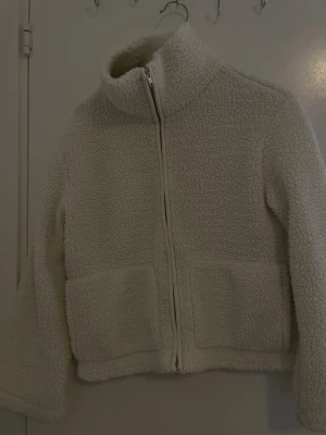 Vit teddyjacka med dragkedja - Mysig vit teddyjacka i fluffigt material med hög krage och hel dragkedja framtill. Jackan har två stora fickor på framsidan och en avslappnad passform. Perfekt för kalla dagar när du vill vara både varm och stilren.