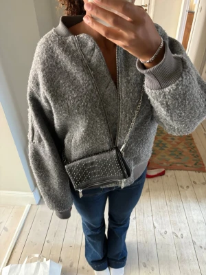 Svart handväska med nitar och kedja - Snygg svart handväska i konstläder med silverfärgade nitar på framsidan och en cool kedjerem. Väskan har dragkedja nertill och är perfekt för att bära det viktigaste. Passar dig som gillar en edgy och trendig stil. Aldrig använd!