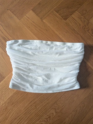 Vit bandeau tubtopp med rynk - Säljer en vit bandeau tubtopp med snygga rynkade detaljer. Toppen är axelbandslös och har en stretchig passform som sitter bekvämt. Perfekt till sommarens outfits och enkel att matcha med olika stilar.