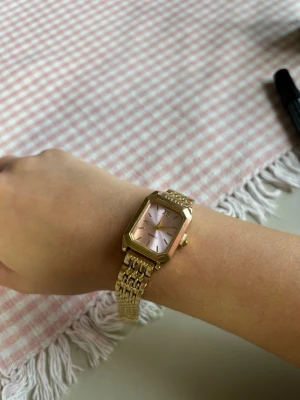Guldig klocka med rosa urtavla - Justerbar storlek medföljer verktyg. Stilren klocka med fyrkantigt guldigt armband och boett. Urtavlan är ljusrosa med guldiga visare och index. Klockan har en elegant och modern känsla, perfekt för dig som gillar accessoarer som sticker ut.