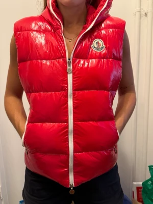 Röd dunväst från Moncler med huva - Snygg röd dunväst från Moncler med glansig finish, vit dragkedja och två fickor framtill. Västen har en huva och Moncler-logga på bröstet. Perfekt för lager-på-lager och streetstyle. KONTAKTA INNAN KÖP