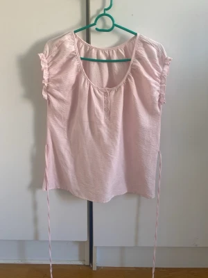 Ljusrosa blus med volangärmar - Supersöt ljusrosa blus med korta volangärmar och knappar framtill. Blusen har rund halsringning och knytband i midjan som ger en fin siluett. Tillverkad i ett mjukt och lätt tyg, perfekt för varma dagar.