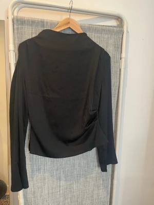 Svart satinblus med lång ärm - Stilren svart blus i ett glansigt satinliknande material. Från H&M i storlek S. Dragkedja i ryggen