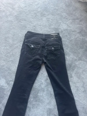 Svarta True Religion jeans - Säljer ett par svarta jeans från True Religion med raka ben och snygga fickdetaljer bak. Jeansen har dekorativa knappar på ficklocken och klassisk True Religion-lapp i midjan. Perfekta för dig som gillar streetwear och vill ha en cool look.