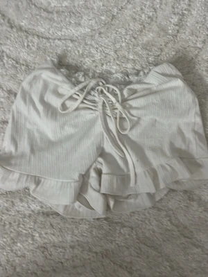 Vita pyjamasshorts med snörning från SHEIN - Söta vita pyjamasshorts med snörning framtill och volangkant. Tillverkade i ett mjukt, ribbat material som känns bekvämt mot huden. Perfekta för mysiga hemmakvällar eller sommarens varma nätter.men den har en liten fläck 