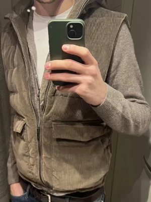 Stenströms Väst - Snygg brun dunväst i manchestertyg med huva och dragkedja framtill. Västen har två stora fickor med lock och quiltad design som ger en modern look. Perfekt att slänga över en hoodie eller tröja för extra värme och stil.