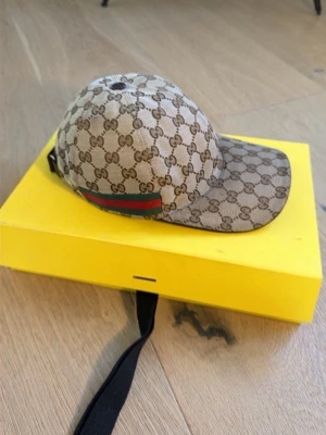 ÄKTA Gucci beige keps med GG-mönster - Snygg beige keps från Gucci med klassiskt GG-monogram över hela kepsen och en grön-röd rand på sidan. Kepsen är i storlek M (58 cm) och har justerbart band bak. Tillverkad i canvasmaterial med svart insida. KVITTO FINNS OCH INGÅR