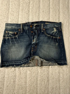 Blå jeanskjol med slitningar - Kort jeanskjol i blå denim med rå kant och slitna detaljer både fram och bak. Kjolen har klassiska fem fickor, bälteshällor och knäppning med knappar framtill. Perfekt för en avslappnad och trendig look. Mjemått är 37cm