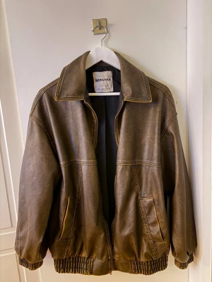 Brun skinnjacka från Bershka - Snygg brun skinnjacka från Bershka med oversized passform och bred krage. Jackan har två framfickor, markerade sömmar och resår vid midja och ärmslut för en cool look. Perfekt för dig som vill ha en trendig och avslappnad stil.