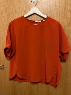 Orange kortärmad topp från Uniqlo - Snygg orange topp från Uniqlo med rund halsringning och avslappnad passform. Toppen har korta ärmar med uppvik och är tillverkad i ett mjukt, lätt material som ger en skön känsla. Perfekt att styla med jeans eller kjol för en trendig look.