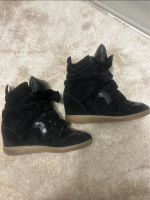 Svarta isabelmarant - Säljer ett par svarta sneakers med kilklack och kardborreband. Skorna har en cool mix av mocka och glansiga detaljer samt en brun sula. Perfekta för dig som vill ha lite extra höjd men ändå sneakers-vibe.