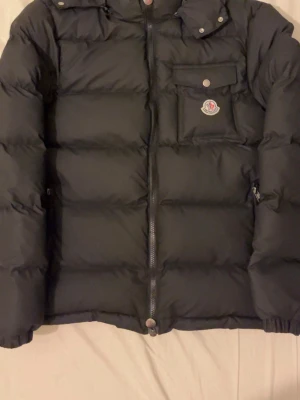 Svart dunjacka från Moncler - Säljer en svart dunjacka från Moncler med klassisk puffad design och hög krage. Jackan har dragkedja framtill, en bröstficka med knapp och Moncler-logga på bröstet. Perfekt för kalla vinterdagar och riktigt snygg till streetwear-stilen.