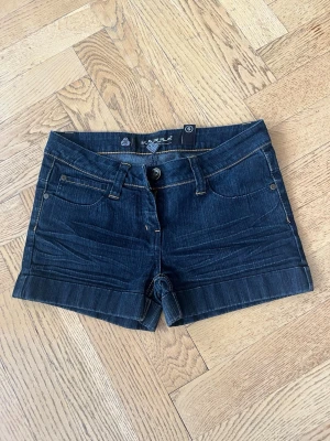 Mörkblå jeansshorts från Karins - Snygga mörkblå jeansshorts från Karins med klassisk femficksdesign, bälteshällor och kontrastsömmar. Shortsen har en midwaist passform och är tillverkade i bomull med en liten uppvikt kant nedtill. Köpta i Spanien!