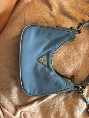 Blå axelremsväska från Guess - Snygg blå handväska från Guess med klassisk trekantslogga i guld framtill. Väskan har en mjuk, rundad form och justerbar axelrem. Stängs med dragkedja. Använd 1 gång