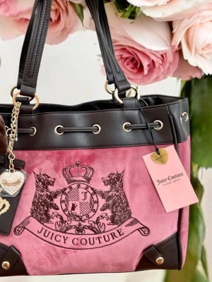 Juicy Couture rosa och svart handväska - Stilren handväska från Juicy Couture i rosa sammet med svarta detaljer i konstläder. Väskan har två handtag, guldfärgade metalldetaljer, dekorativ rosett och emblem med lejon framtill. Hängande hjärta och tag med logga ingår.