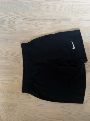 Svarta träningsshorts från Nike - Säljer ett par svarta träningsshorts från Nike med klassisk vit logga på benet. Shortsen har elastisk midja och är tillverkade i ett lätt och bekvämt material som passar perfekt för sport och aktivitet.