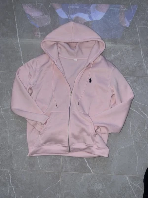 Ljusrosa hoodie med dragkedja - Säljer en ljusrosa hoodie med dragkedja och huva. Hoodien har fickor framtill och en broderad logga på bröstet. Tillverkad i mjukt material och har justerbara snören i huvan. Perfekt för en chill och avslappnad stil.