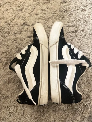 Svarta och vita Vans Old Skool sneakers - Klassiska Vans Old Skool sneakers i svart och vitt med den ikoniska vita sidan, vita snören och vit sula. Ovandelen är i mocka och canvas, vilket ger en snygg kontrast. Perfekta för en avslappnad streetwear-look. Skorna har använts i cirka 6 månader och som man kan se på bilden så finns de spår av användning