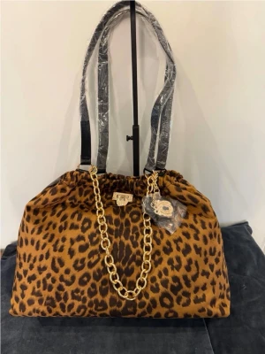 Juicy Couture Leopardmönstrad handväska viral  - Snygg handväska i leopardmönstrat tyg med detaljer i guld, inklusive en kraftig guldfärgad kedja och dekorativt hängsmycke. Väskan har svarta axelband och stängs med ett guldfärgat lås framtill. Perfekt för dig som vill sticka ut med ett djurmönster.