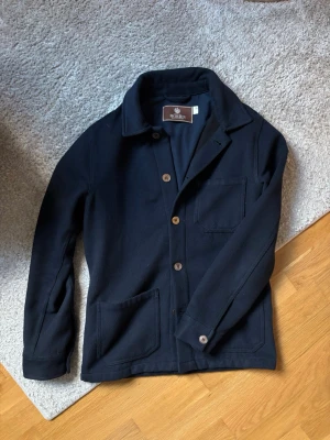 Mörkblå overshirt från Morris - Stilren mörkblå overshirt från Morris med klassisk krage och knäppning framtill. Tre fickor, varav en bröstficka och två framtill. Overshirten har en rak passform och snygga bruna knappar. Perfekt lager på lager-plagg för en trendig look.