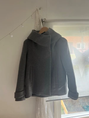 Grå ullkappa med stor krage - Snygg grå kappa i ullmix med bred, asymmetrisk krage och raka fickor framtill. Kappan har en modern, boxig passform och uppvikta ärmslut som ger en trendig look. Perfekt för kyliga dagar när du vill vara både varm och stilren.