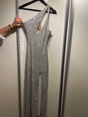 Glittrig silver one shoulder-klänning - Snygg lång klänning i silver med massor av paljetter som ger ett riktigt glittrigt intryck. One shoulder-modell med asymmetrisk skärning och figurnära passform. Man får med en fluga och en ”näsduk” som matchar, klänningen är från sellpy!! 
