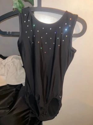 Svart gymnastikdräkt med strass - Svart ärmlös gymnastikdräkt i stretchigt material med rund halsringning. Framsidan är dekorerad med små glittrande strasstenar som ger en snygg look. Perfekt för träning inom gymnastik eller dans.