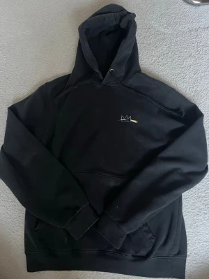 Hov1 hoodie - Säljer en svart hoodie med huva och dragsnören. På bröstet finns ett litet, minimalistiskt vitt tryck av en krona med en gul linje. Hoodien har känguruficka framtill och ribbade muddar vid ärmar och nederkant. Perfekt för en avslappnad streetwear-look.