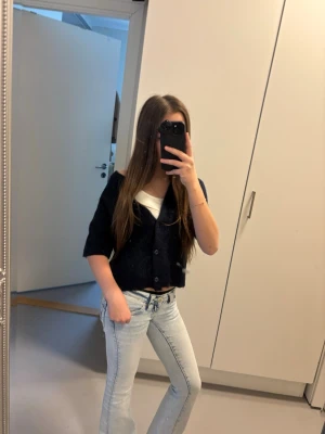 Svart kofta från Gina Tricot - Mysig svart kofta från Gina Tricot med knappar framtill och ribbade muddar. Kortare modell som passar perfekt till jeans eller kjol. Stickad i mjukt material och har en enkel, stilren look.