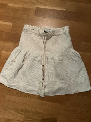 Vit/beige kortkjol med knyt och volang - Supersöt vit/beige kortkjol med smock i midjan, knytband med tofsar och små pärlor framtill. Kjolen har volang och ett diskret strukturerat mönster. Kjolen kan i vissa ljus se något flammig ut!😊