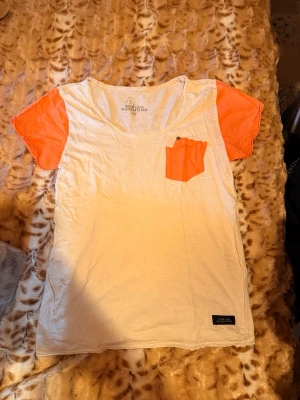 Vit t-shirt med orange detaljer Adrian hammond - Säljer en vit t-shirt med korta orange ärmar och matchande orange bröstficka. T-shirten har en avslappnad passform och är tillverkad i mjuk bomull, perfekt för en chill och stilren look.